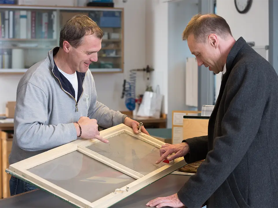 Glasermeister Günther Reisch im Gespräch mit einem Kunden einen Fensterflügel haltend